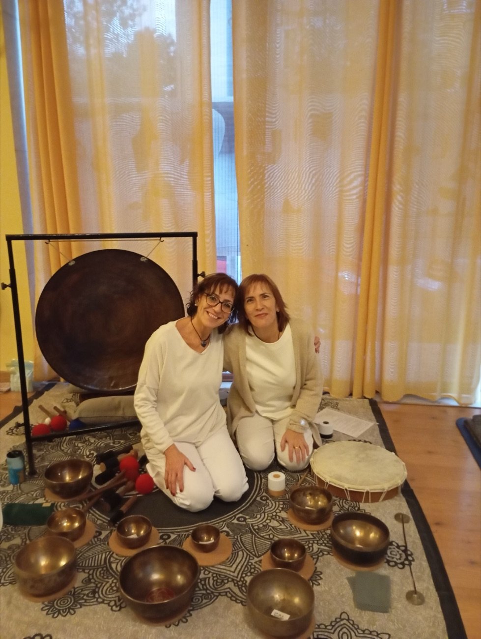 Maribel y Sonia, vestidas de blanco, sentadas junto al gong y los cuencos en una sesión de baño de sonido.